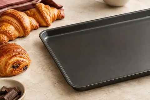 Plaque pâtissière rectangulaire noire en aluminium antiadhésif près de viennoiseries sur un plan clair.