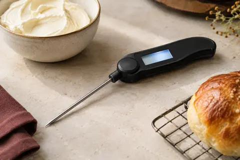 Thermomètre de cuisine noir pliable, sonde déployée près d’un pain et d’un bol de crème
