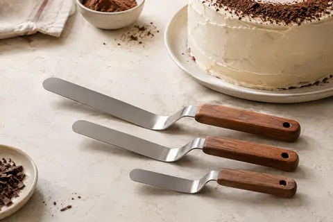 Trois spatules coudées en inox à manches en bois posées près d’un gâteau glacé.