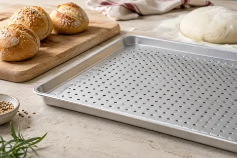 Plaque de cuisson perforée en aluminium argenté posée près de petits pains dorés sur un plan clair.