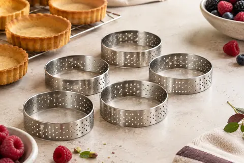 Six cercles à tartelettes perforés en inox posés près de petits fonds de pâte dorés
