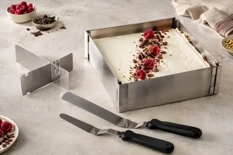 Cadre pâtissier extensible en inox avec séparateur et spatules noires près d’un gâteau à glacer.
