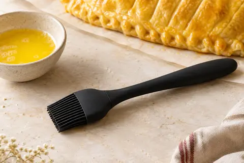 Pinceau de pâtisserie noir en silicone posé près d’une pâte dorée et d’un bol de beurre fondu
