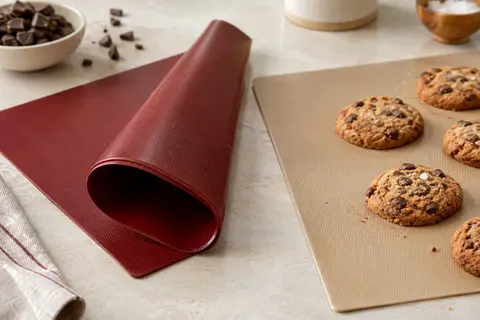 Deux tapis de cuisson en silicone rouge et beige posés sur un plan de travail en pierre claire, près de biscuits.